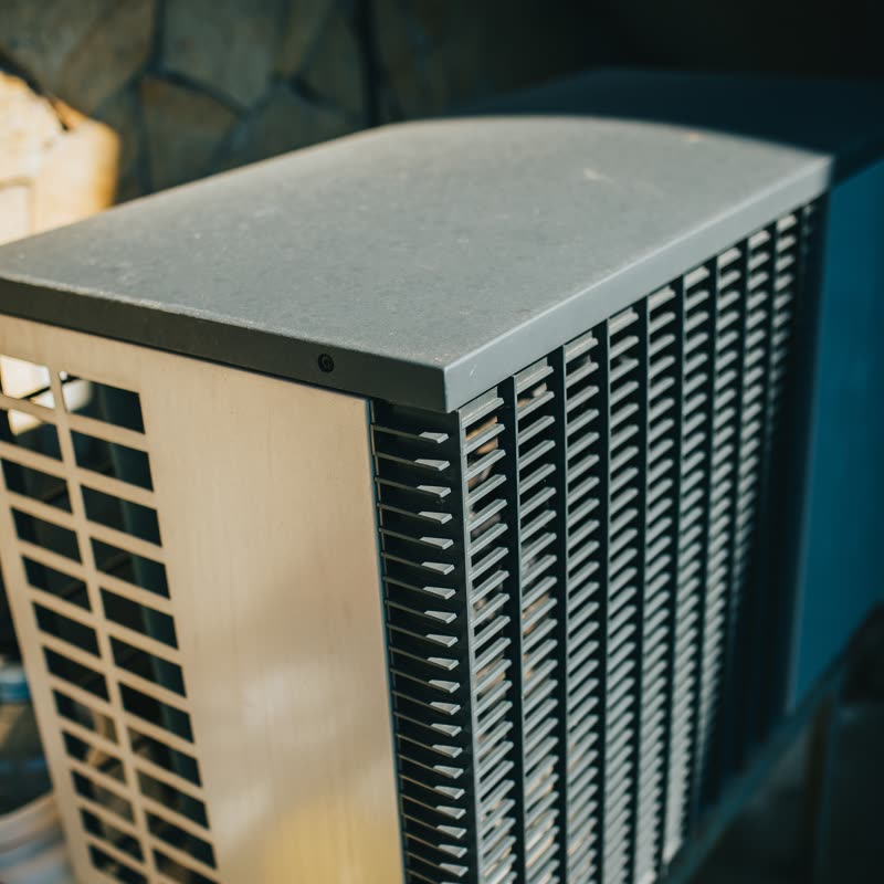 Late sun across the side panel and fins of an outdoor air conditioning unit beside a stone garden wall